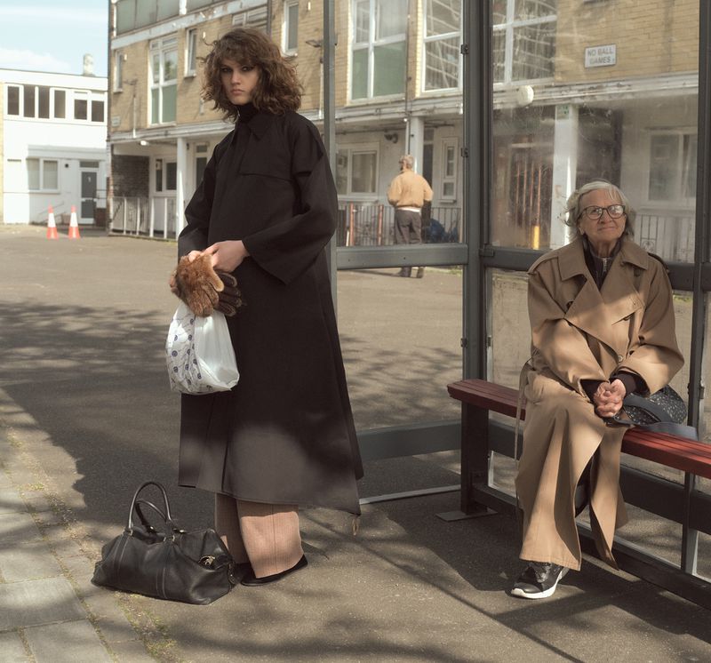 Model standing by a bus stop wearing a black coat while an older woman sits on the bench