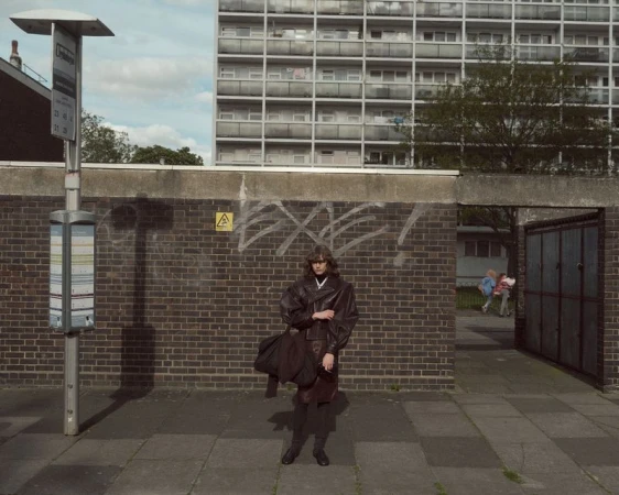Model standing on a paved street in front of a brick wall and bus stop wearing a dark leather jacket holding a dark bag