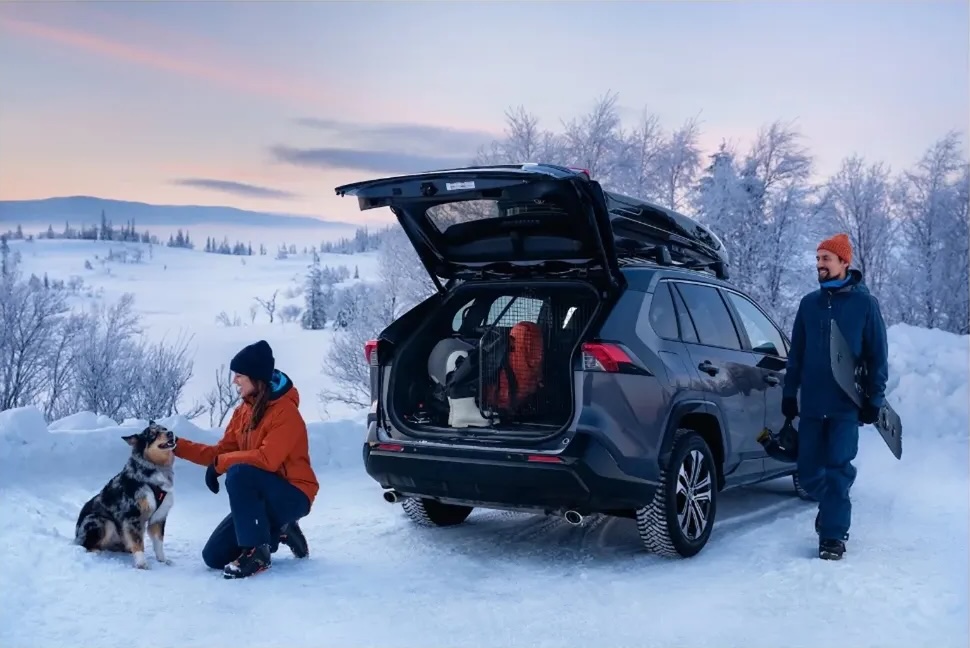Woman pets dog near SUV with snowy mountain backdrop, while man holds snowboard. Winter scene with vehicle, people, animal, and nature.