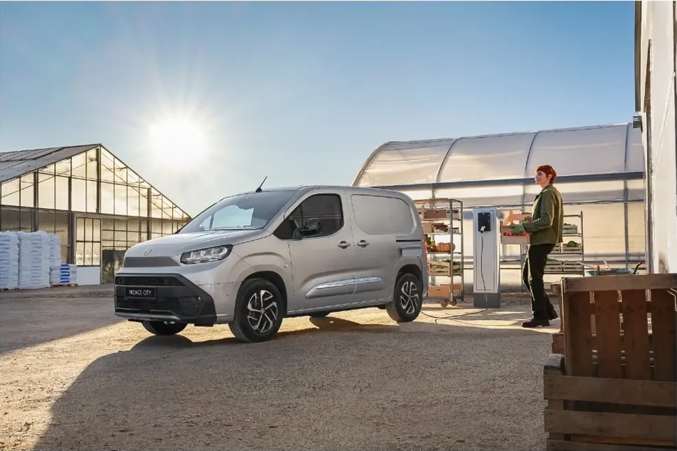 Silver commercial van parked near greenhouses, with an adult carrying a crate, and electric vehicle charging station in daytime.