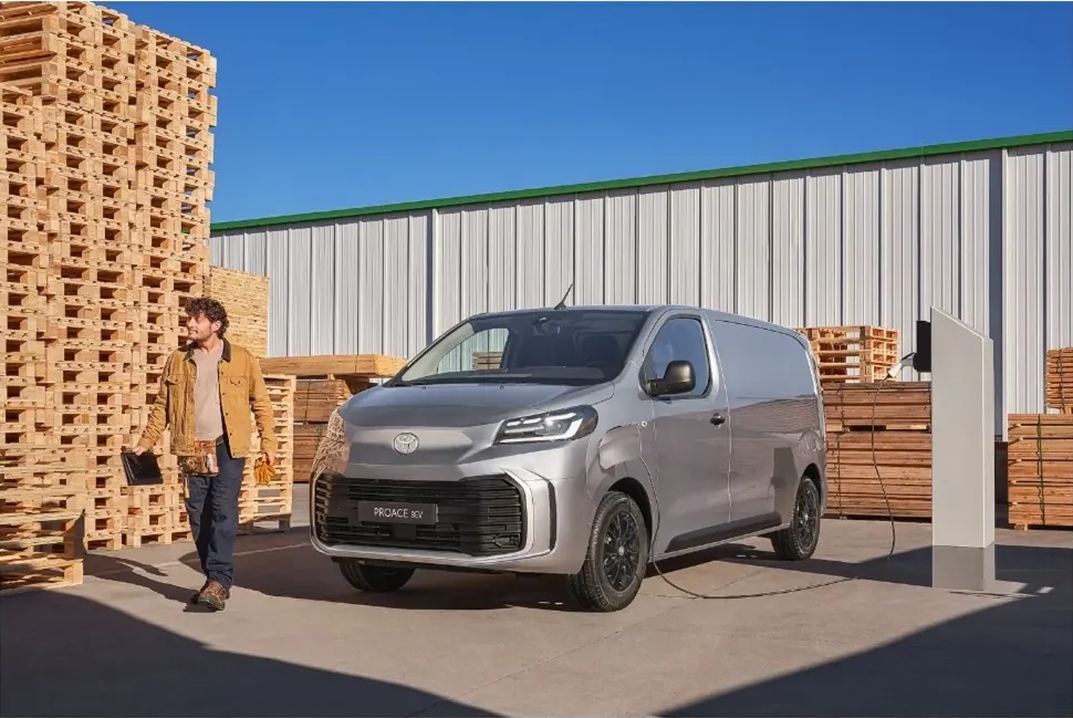 Silver electric delivery van parked outdoors with a man in a tan jacket walking nearby, wooden pallets and corrugated metal in the background.