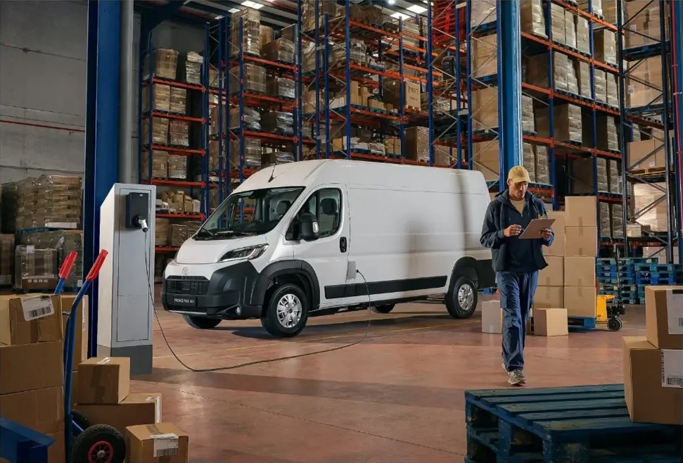 White electric cargo van charging indoors in a large warehouse, with a worker holding a tablet near stacked boxes and metal shelves.