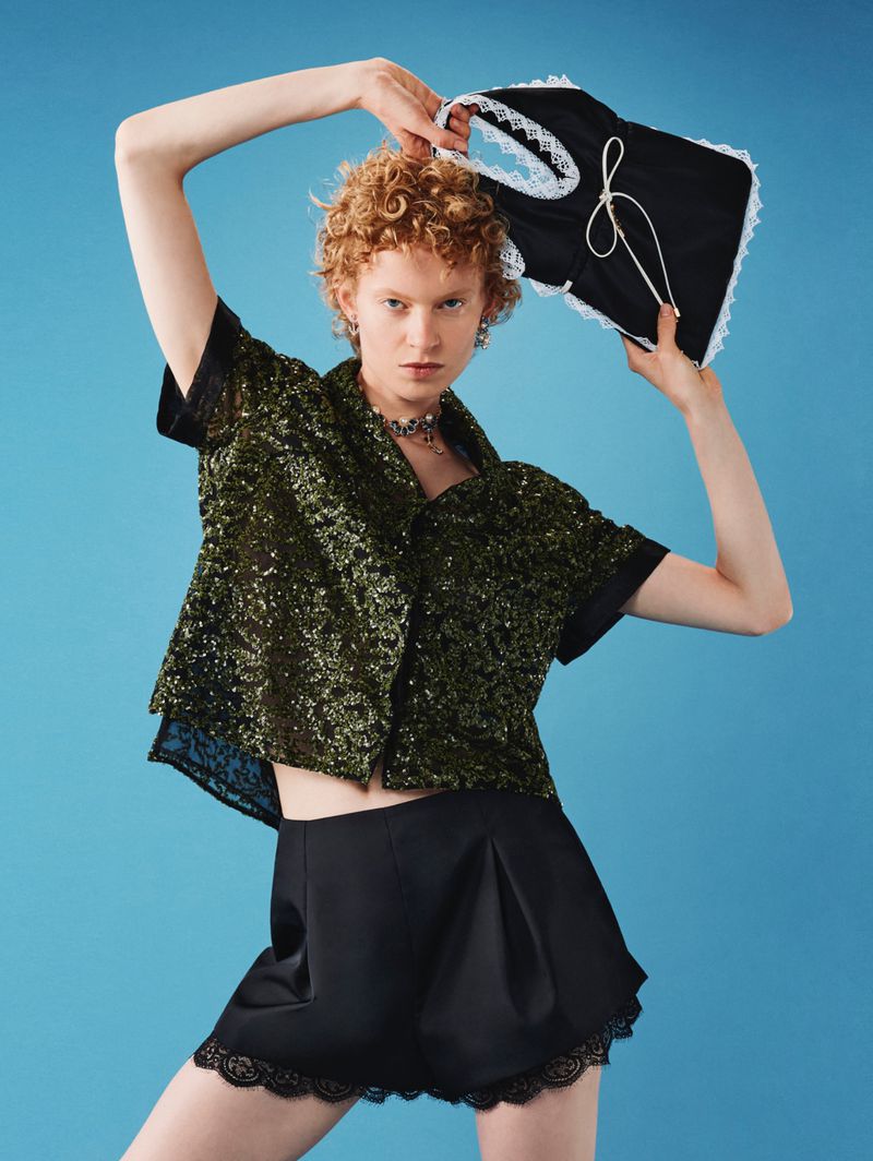 Young woman in a shimmering olive top and black lace-trimmed shorts holds a black and white garment in a studio shot against blue.