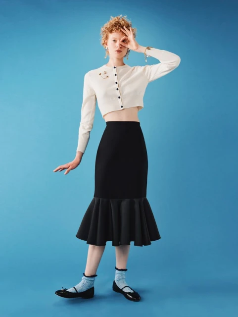 Fashion shot of a woman in a white cardigan and black skirt posing against a blue background, showing full-body studio portrait.
