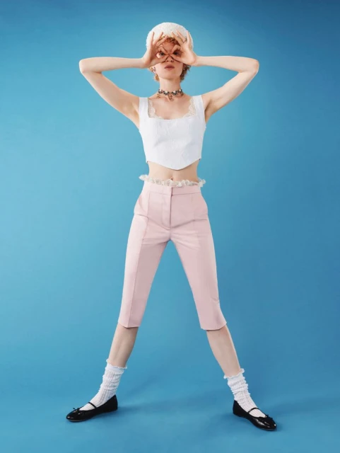 Full shot of a young woman in a white top and pink capri pants making circles around her eyes with her hands against a blue background.