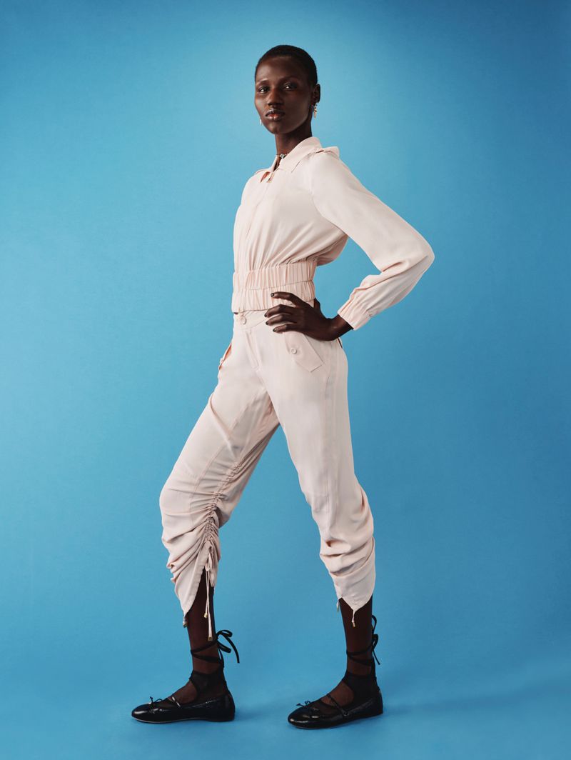 Fashionable woman in studio with a modern pale pink top and pants with black flats poses against a blue backdrop.