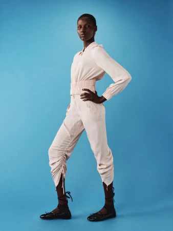 Fashionable woman in studio with a modern pale pink top and pants with black flats poses against a blue backdrop.