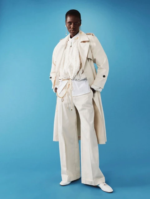 Fashionable black model poses in beige trench coat, wide-leg pants, and white top on a light blue backdrop in studio lighting.
