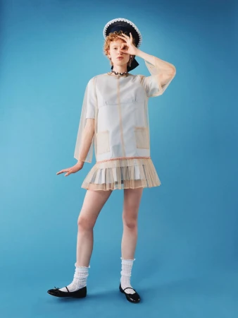 A young woman with curly red hair poses in a sheer white dress with a sailor hat against a vibrant blue background in a studio shot.