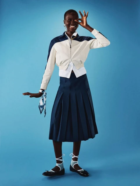 Full-length studio shot of a smiling Black woman in a white jacket, navy pleated skirt, and ballet flats against a light blue background.