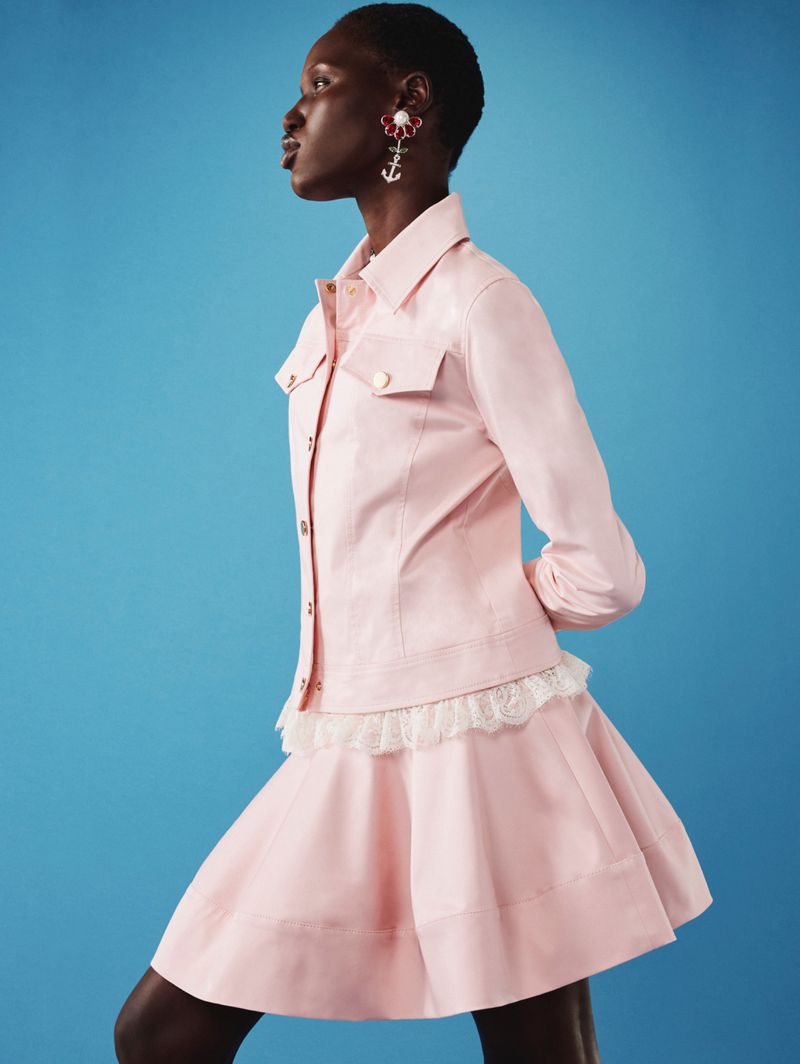 Side profile studio shot of a Black female model in a light pink dress and jacket, posing against a blue backdrop with drop earrings.