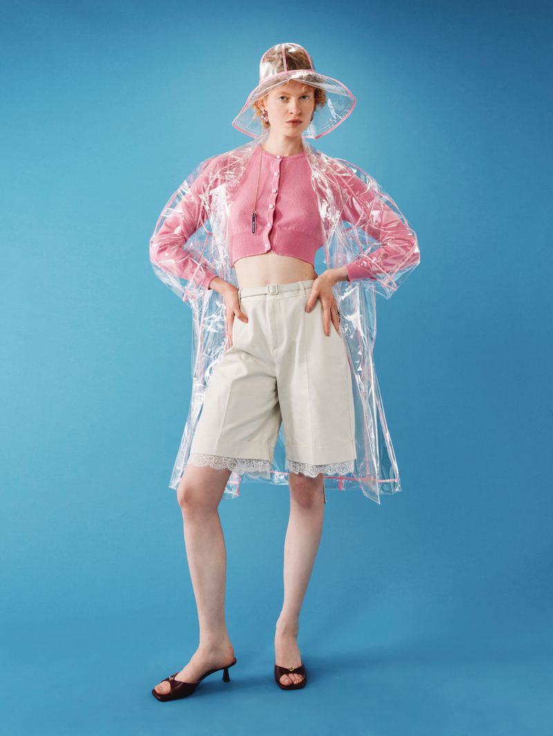 Fashion portrait of a woman wearing a transparent rain hat and coat, pink top, and cream shorts against a solid blue background.