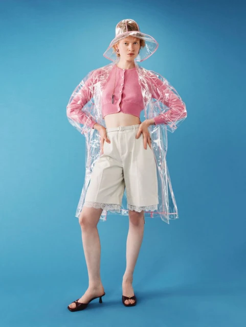 Fashion portrait of a woman wearing a transparent rain hat and coat, pink top, and cream shorts against a solid blue background.