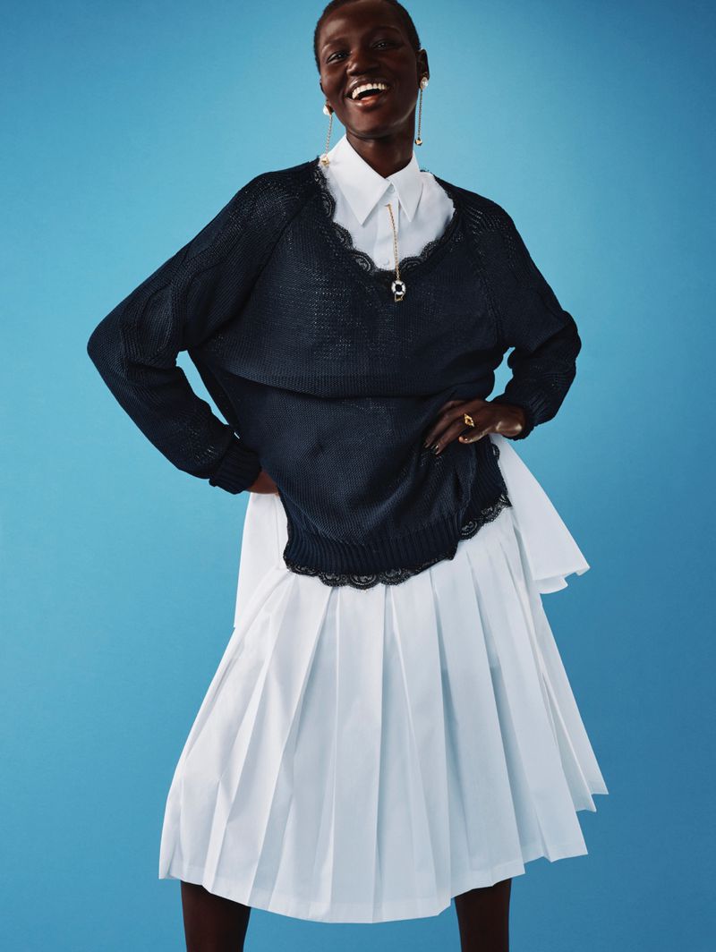 Smiling woman in an elegant, dark blue sweater with lace trim, layered over a white shirt and pleated skirt, against a light blue background.