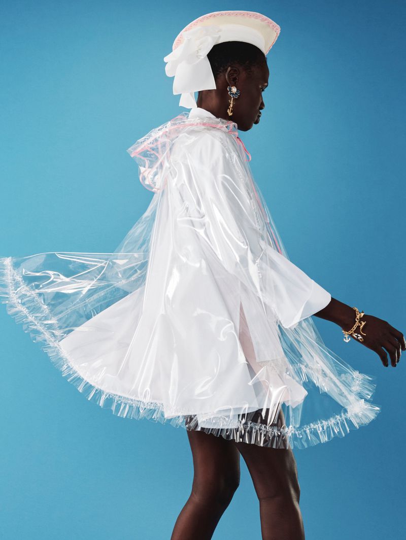 A dark-skinned model in a transparent raincoat and hat poses against a vibrant blue backdrop, showcasing fashion and style.