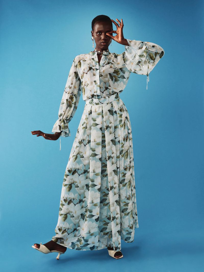 Elegant adult female fashion model poses in long floral chiffon dress against a blue background in a studio, wearing white heels.