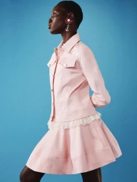 Model Eman Deng wearing a light pink jacket and matching skirt with lace trim against a blue backdrop for Vivetta Resort 2026.