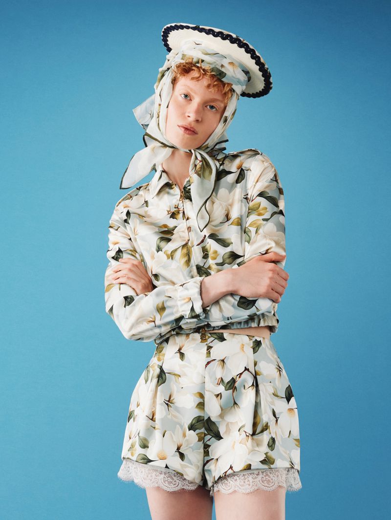 Model wearing a floral jacket, matching lace-trimmed shorts, silk headscarf, and hat against a solid sky blue studio backdrop.