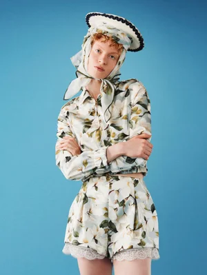 Model wearing a floral jacket, matching lace-trimmed shorts, silk headscarf, and hat against a solid sky blue studio backdrop.