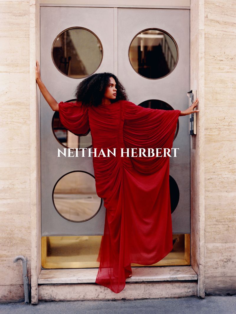 Adult female fashion model in a red gown poses in front of a metal door with circular windows, blending modern elegance and architectural design.