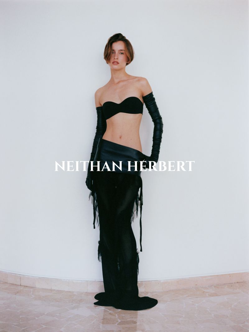 Fashion photo of a slender adult woman in black strapless top, arm gloves, and low-slung pants, posing in front of a white wall.