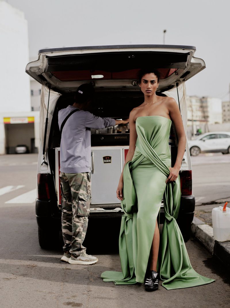 Model Imaan Hammam posing in a draped sage green strapless gown and black sneakers next to an open van on an asphalt lot.