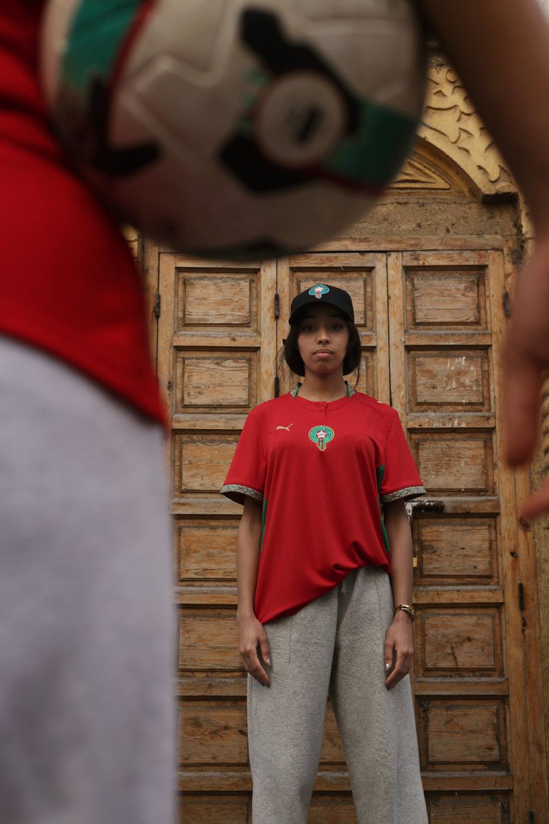 Model Imaan Hammam wearing a red soccer jersey, grey sweatpants, and a black cap framed behind an out-of-focus soccer ball.