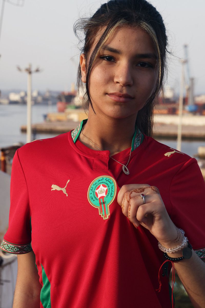 Young woman in a red Morocco national soccer jersey holding the team crest against an outdoor port background.