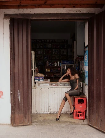Model Imaan Hammam seated on red crates in a dark dress before a rustic shop counter framed by open brown metal doors.