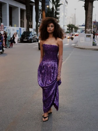 Model Imaan Hammam walks along an urban street wearing a strapless purple sequin gown and opera gloves.