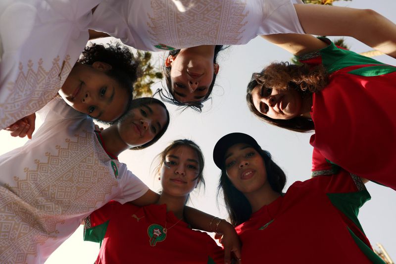 Low angle view of model Imaan Hammam and a group of young women in Moroccan soccer jerseys forming a huddle looking down at camera.