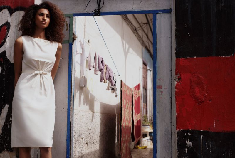 Model Imaan Hammam posed in a sleeveless white belted dress leaning against an open doorway leading to a courtyard with hanging laundry.