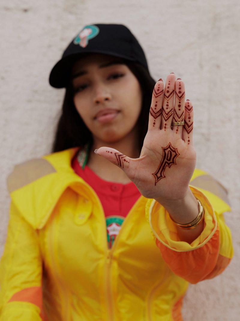 Model Imaan Hammam holding her hand forward showing henna patterns and a P logo, wearing a yellow jacket and black cap.