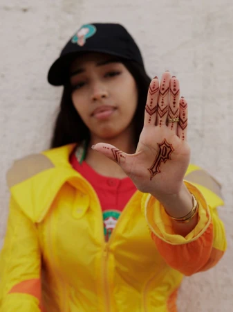 Model Imaan Hammam holding her hand forward showing henna patterns and a P logo, wearing a yellow jacket and black cap.