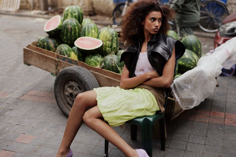 Model Imaan Hammam sitting on a green plastic chair in a leather vest and yellow skirt in front of a cart full of watermelons.