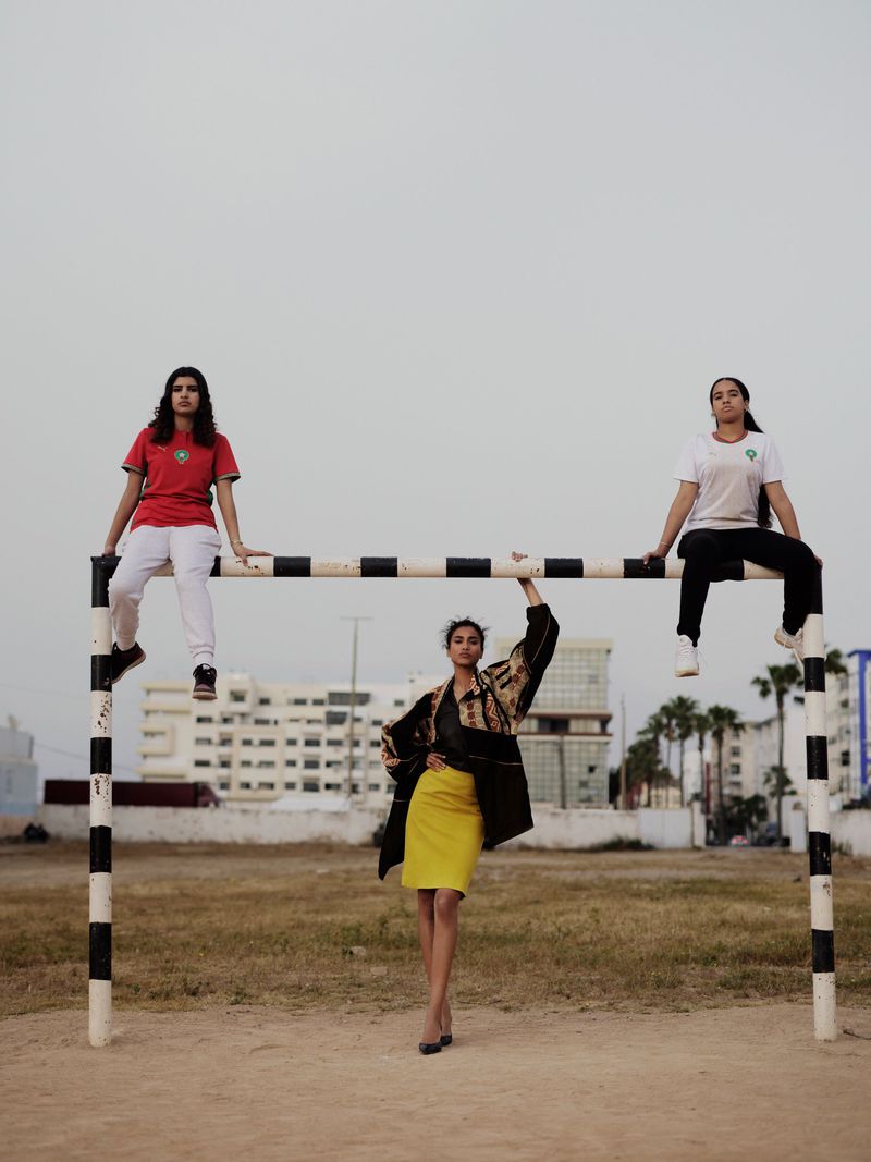 Model Imaan Hammam in a yellow skirt stands under a soccer goalpost beneath two women sitting on the crossbar in athletic jerseys.