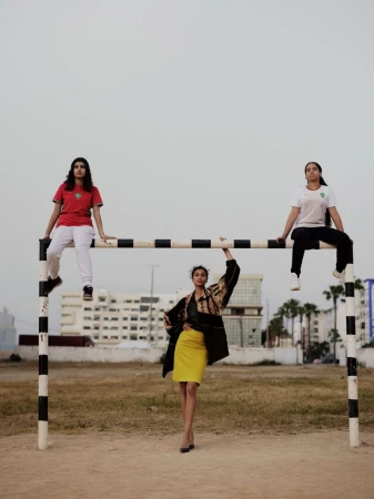 Model Imaan Hammam in a yellow skirt stands under a soccer goalpost beneath two women sitting on the crossbar in athletic jerseys.