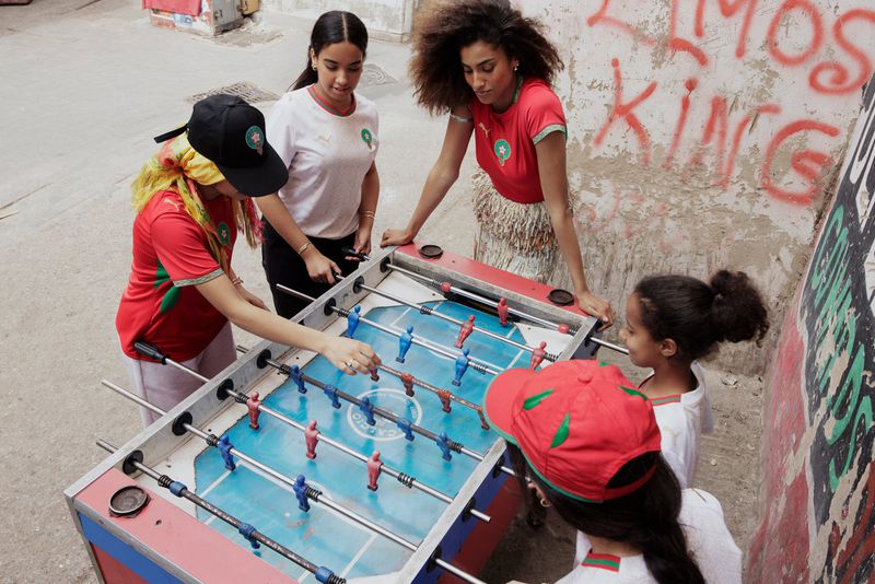 Imaan Hammam and young women in Morocco soccer jerseys gathered around an outdoor foosball table by a graffiti wall.