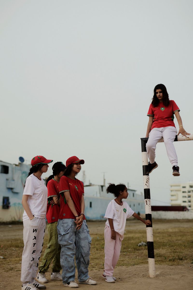 A group of young women wearing red soccer jerseys and casual streetwear on an open dirt field, with one sitting on a goalpost.