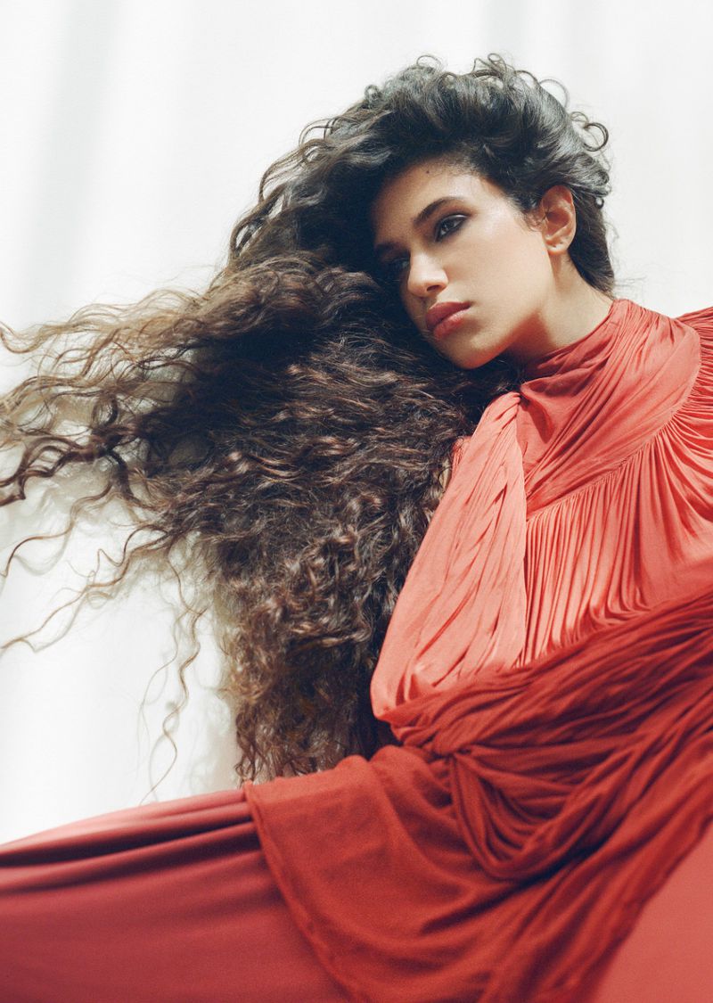 Model Tamar David in a draped pleated coral dress with long voluminous curls against a white backdrop.