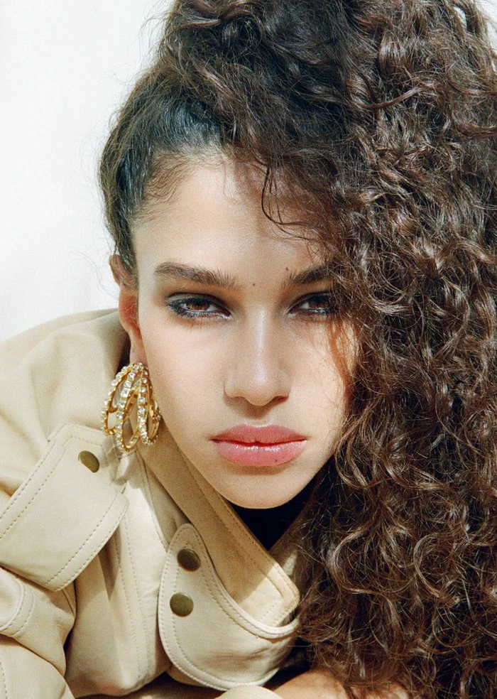 Close-up portrait of model Tamar David wearing large gold hoop earrings and a beige jacket with curly brown hair.