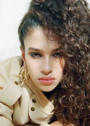 Close-up portrait of model Tamar David wearing large gold hoop earrings and a beige jacket with curly brown hair.