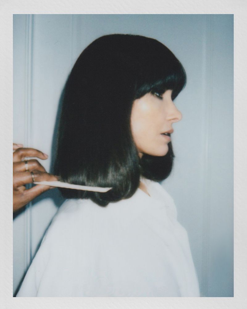 Woman with dark bob haircut getting hair combed by a person in a photo studio with bright lighting and soft focus.