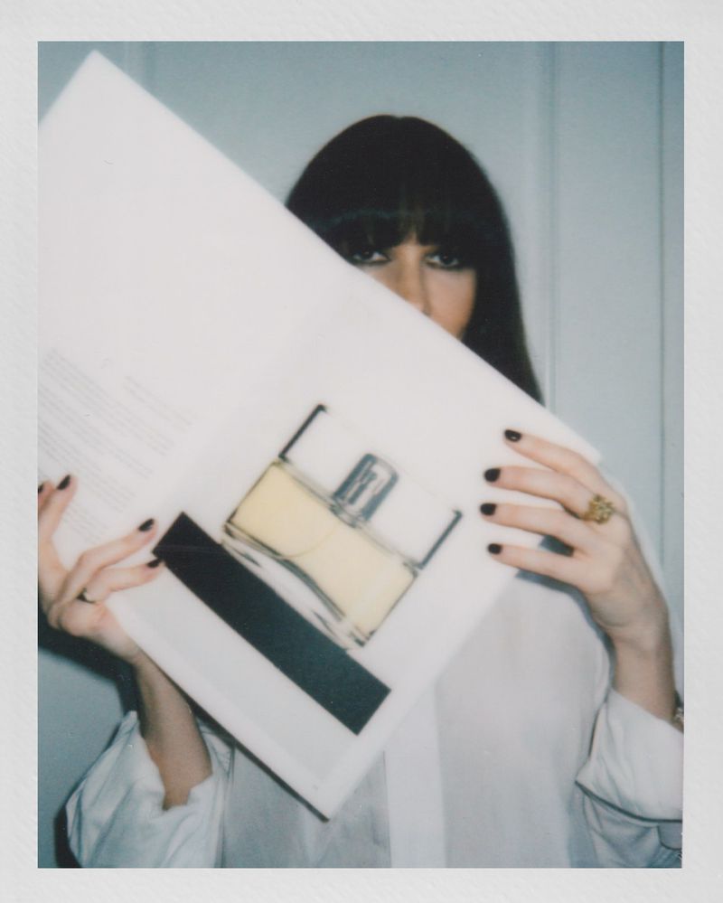 Woman with dark hair holds up a large white portfolio in front of her face, showing a drawing of a yellow perfume bottle.