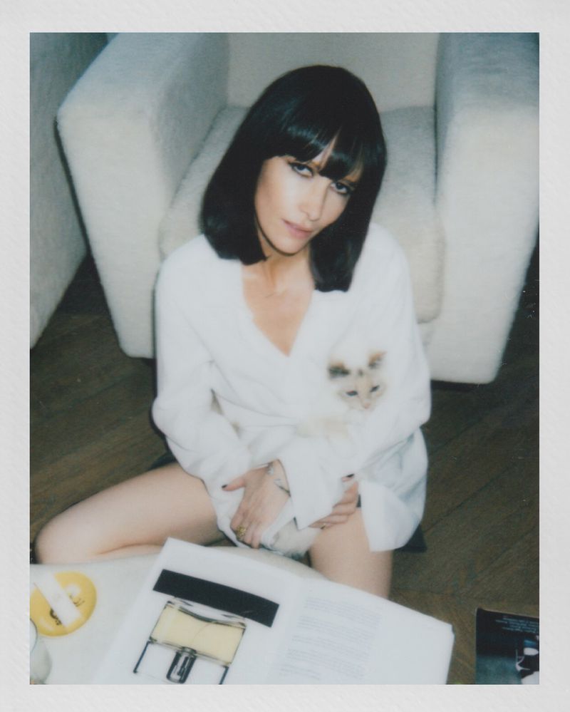 Brunette woman in a white robe poses indoors with a fluffy cat, papers, and a drink for a casual, relaxed portrait.