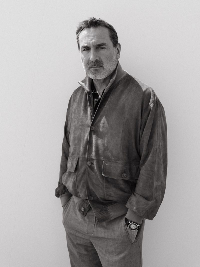 Black and white portrait of a man in a suede jacket and trousers standing with hands in pockets against a plain backdrop.