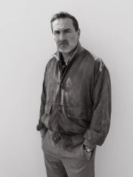 Black and white portrait of a man in a suede jacket and trousers standing with hands in pockets against a plain backdrop.
