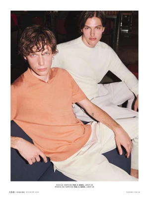 Two male models posing indoors, one in a peach polo shirt and cream pants and the other in an off-white long-sleeve knit top.