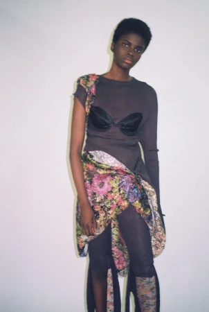 Model Rachel Agbonze posing in a dark knit top with a bra overlay and floral wrapped skirt against a white studio background.