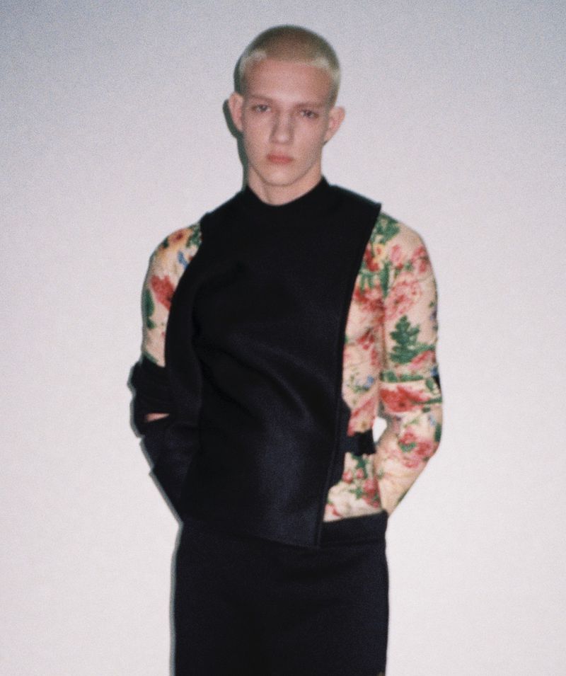 Blonde male model wearing a black structured vest over a floral long-sleeve shirt against a plain light studio backdrop.